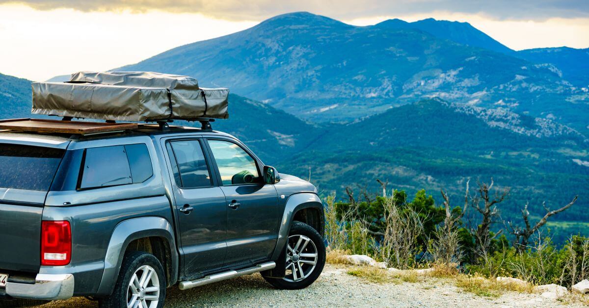 A dark gray truck parked on a hillscape overlooking a mountain range covered with dark green trees and a cloudy sky.