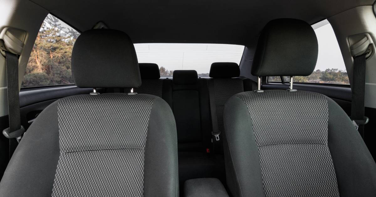 A sedan's interior contains grey upholstery. A forest is visible outside of the vehicle's side and rear windows.