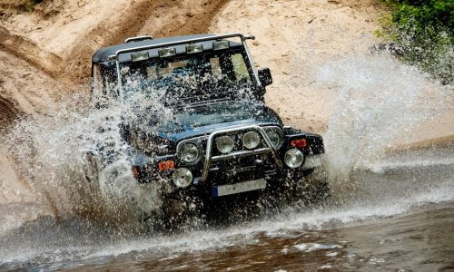 A boxy black Jeep driving down a hill and into a large, muddy puddle with water splashing everywhere.