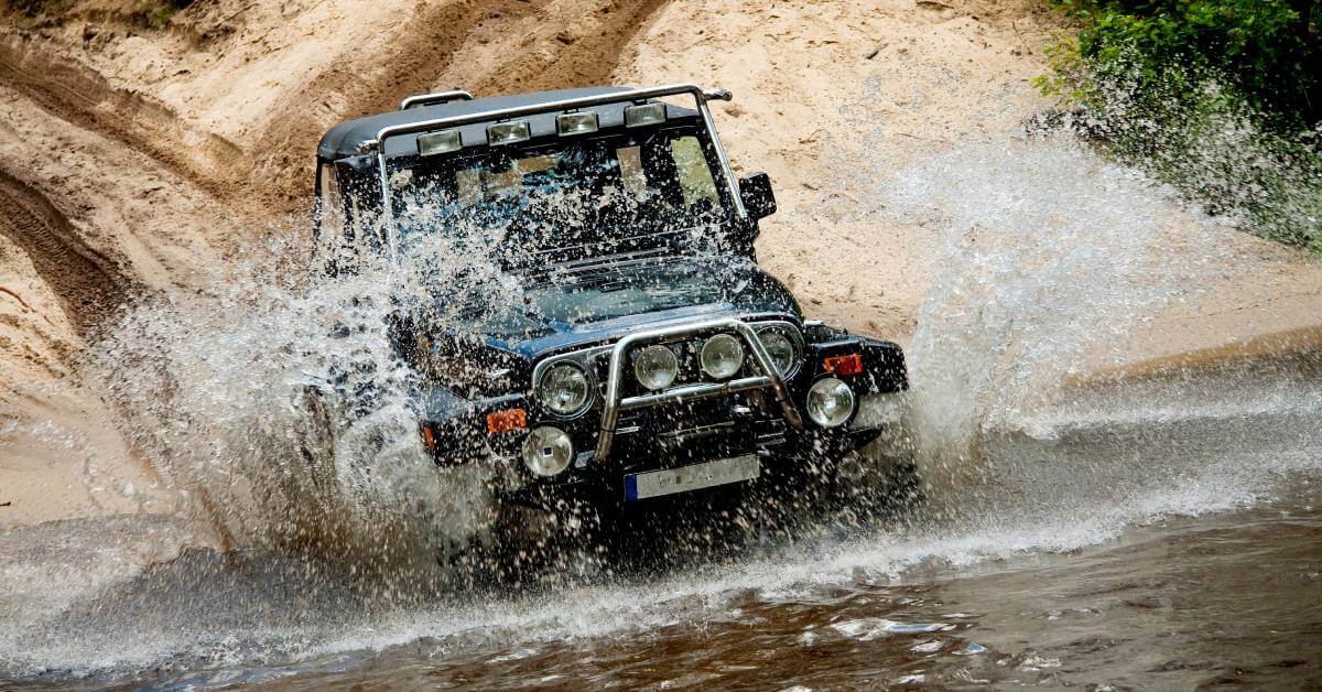 A boxy black Jeep driving down a hill and into a large, muddy puddle with water splashing everywhere.