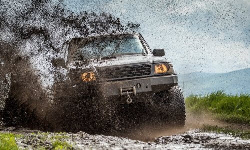 A boxy truck with large tires and a winch at the front driving down a muddy road in the mountains. Mud is sprayed everywhere.