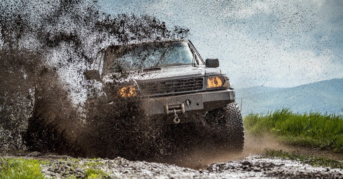 A boxy truck with large tires and a winch at the front driving down a muddy road in the mountains. Mud is sprayed everywhere.