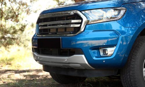 The front and driver's side of a shiny blue pickup truck parked on grass with trees around. Its grill and lights are clean.