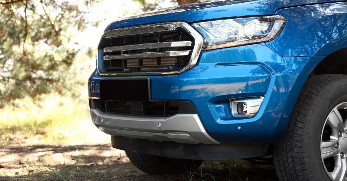 The front and driver's side of a shiny blue pickup truck parked on grass with trees around. Its grill and lights are clean.