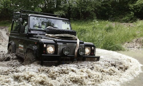 An off-roading vehicle with an extra tire on the hood and a rack on the roof driving through a large muddy puddle in a forest.