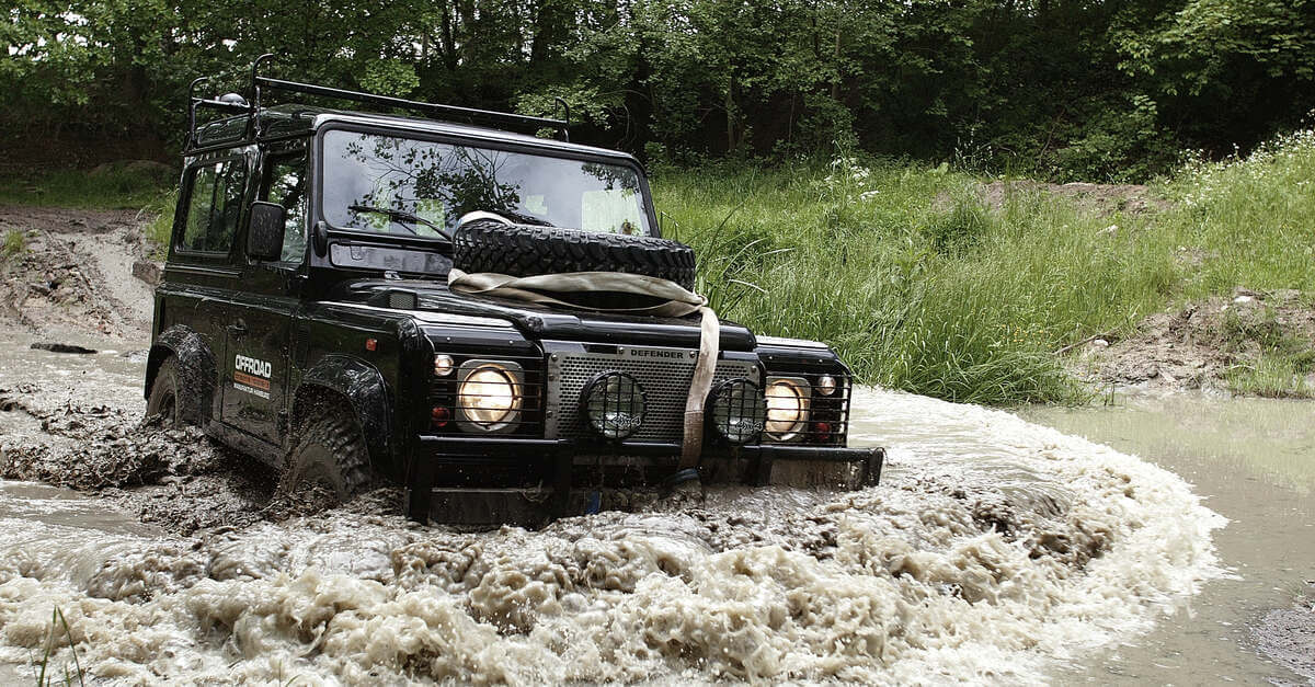 An off-roading vehicle with an extra tire on the hood and a rack on the roof driving through a large muddy puddle in a forest.