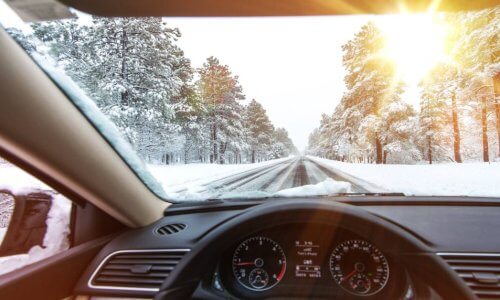 The view from the driver's seat of a car, looking out over a snowy road with trees on either side and sun streaming in.