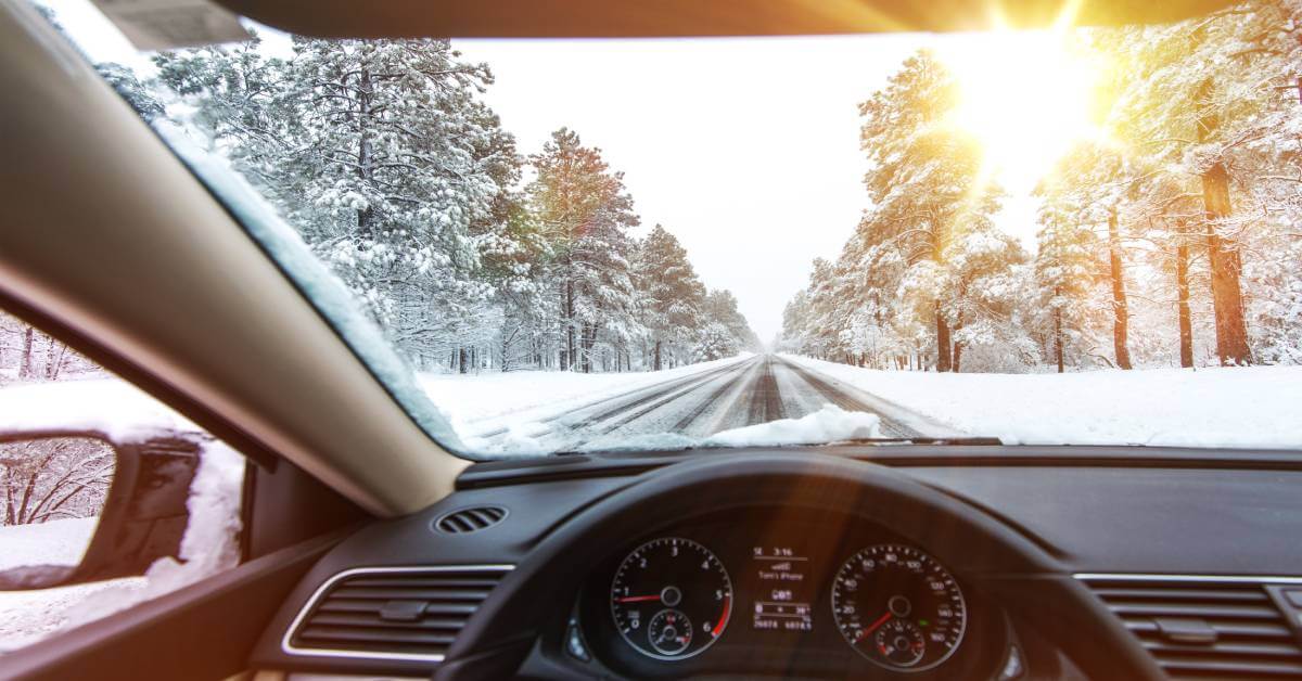 The view from the driver's seat of a car, looking out over a snowy road with trees on either side and sun streaming in.