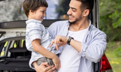 A smiling man plays with a child in his arms. Behind him are trees and a car with its trunk door open.