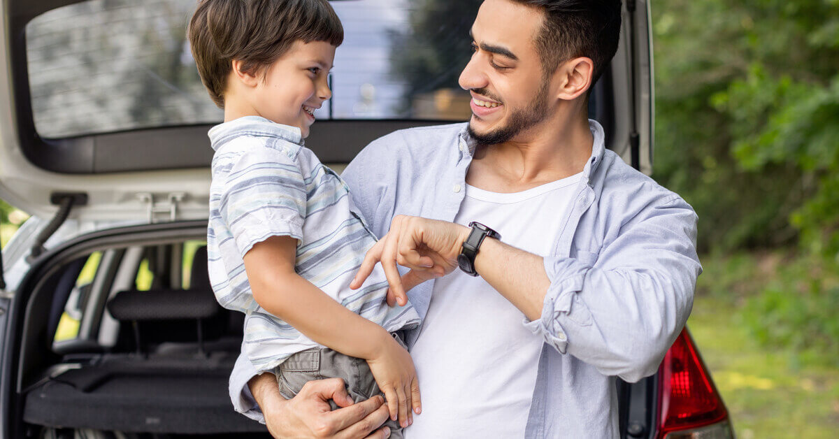 A smiling man plays with a child in his arms. Behind him are trees and a car with its trunk door open.